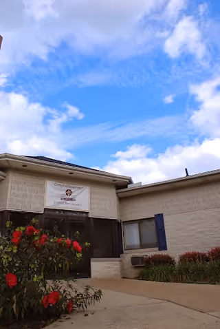 Exterior view of Crestmont North Nursing facility showing a beige brick building with a banner above the entrance, a pathway leading to the door, red flowers and green shrubs in the foreground, and a partly cloudy blue sky.