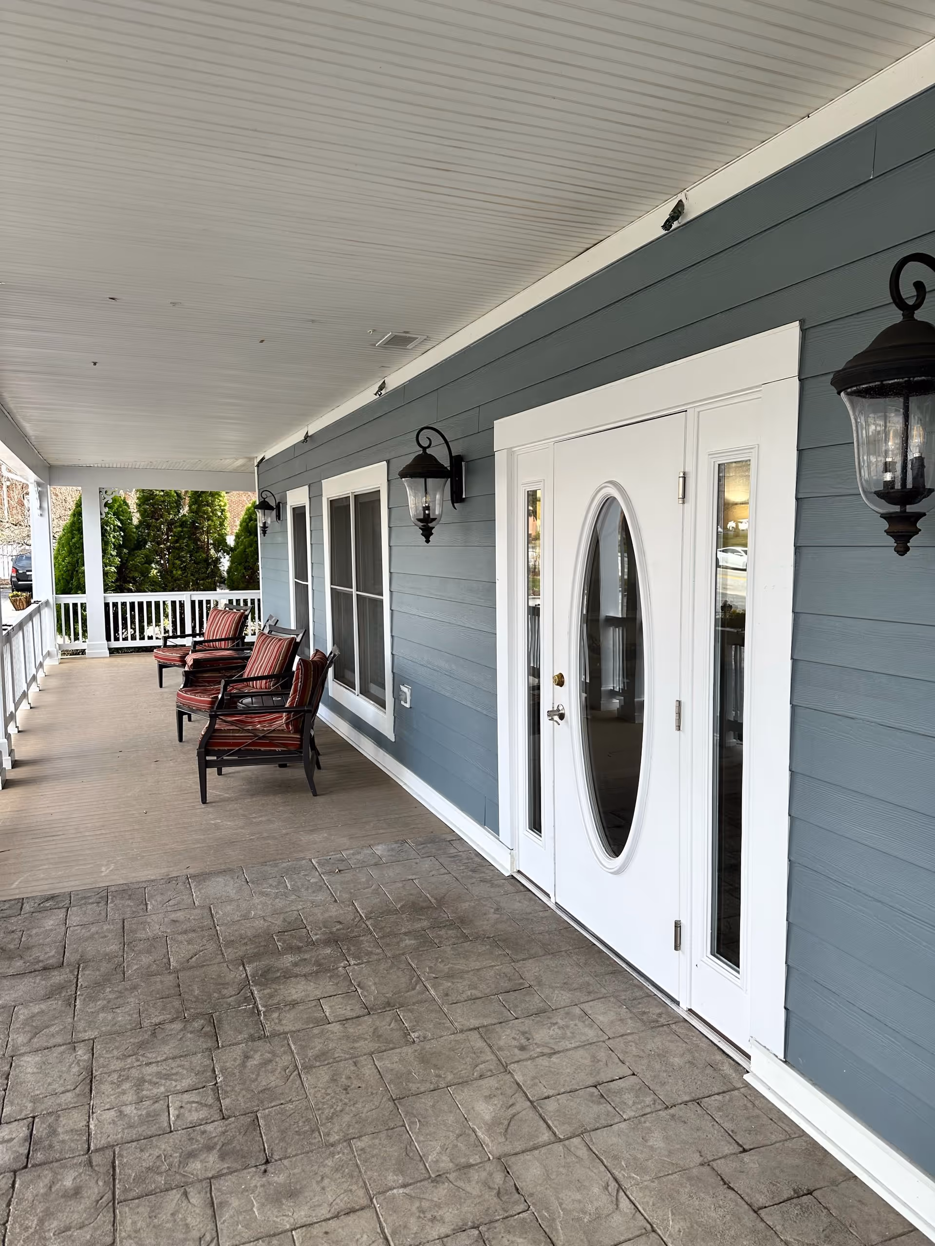 Covered porch area with a white ceiling and blue siding walls featuring two black wall-mounted lanterns. There are three cushioned chairs with red and black striped upholstery arranged along the porch. A white door with an oval glass window and two narrow side windows is visible on the right side. The porch floor is a combination of stamped concrete and wood.