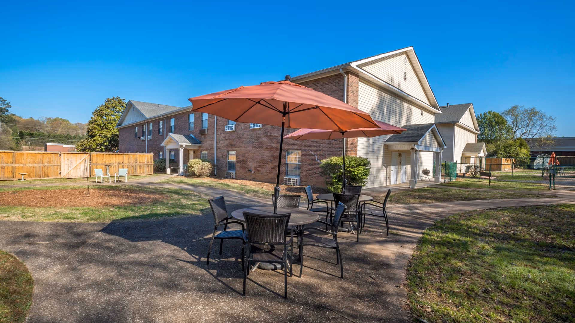 Outdoor patio area with two round tables, each shaded by a large orange umbrella, surrounded by black chairs. The patio is part of a senior living facility with a two-story brick and siding building in the background, a wooden fence, and some greenery under a clear blue sky.