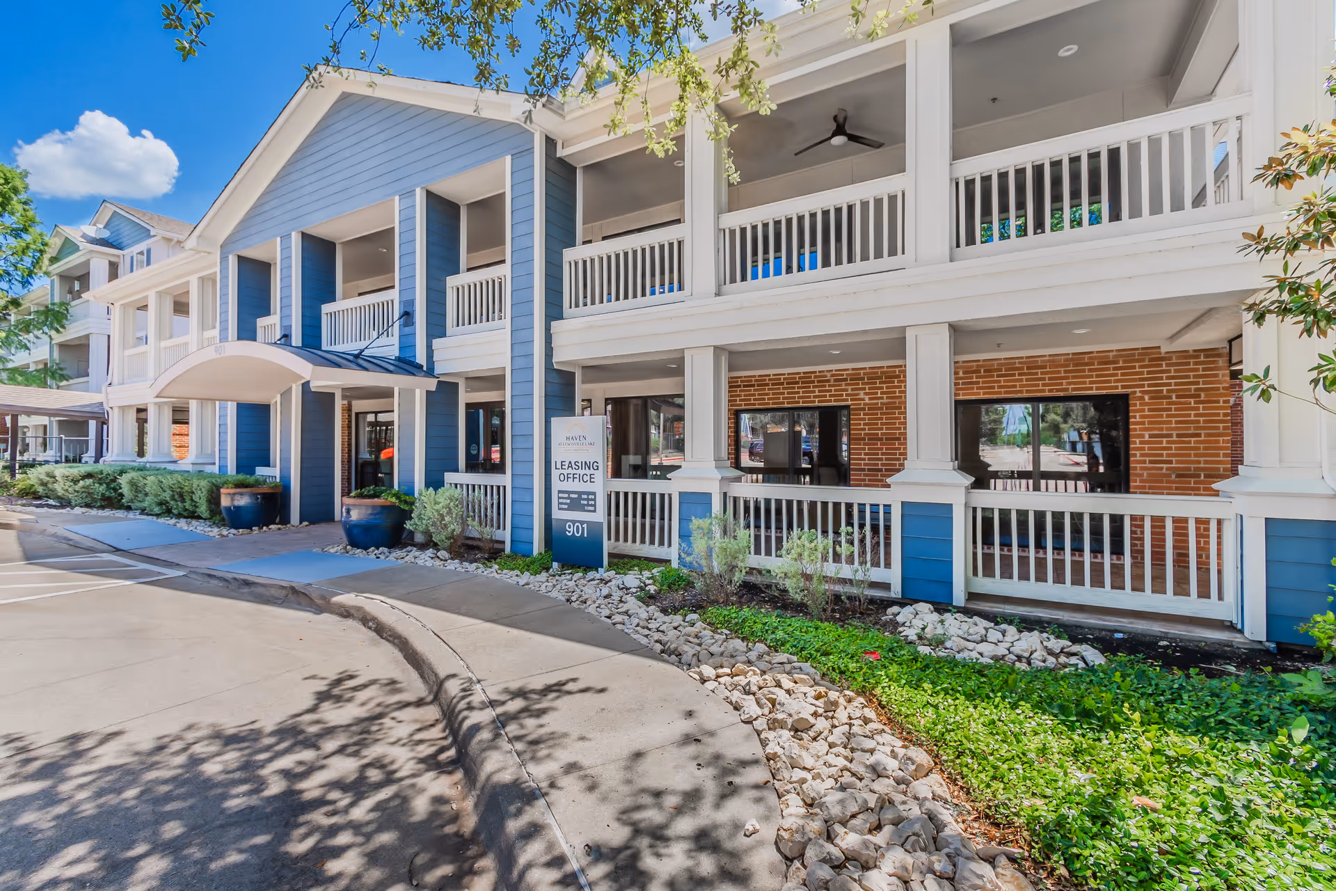 Exterior view of the Haven at Lewisville Lake leasing office building with blue and white siding, brick accents, a covered entrance, and landscaping with rocks and greenery under a clear blue sky.
