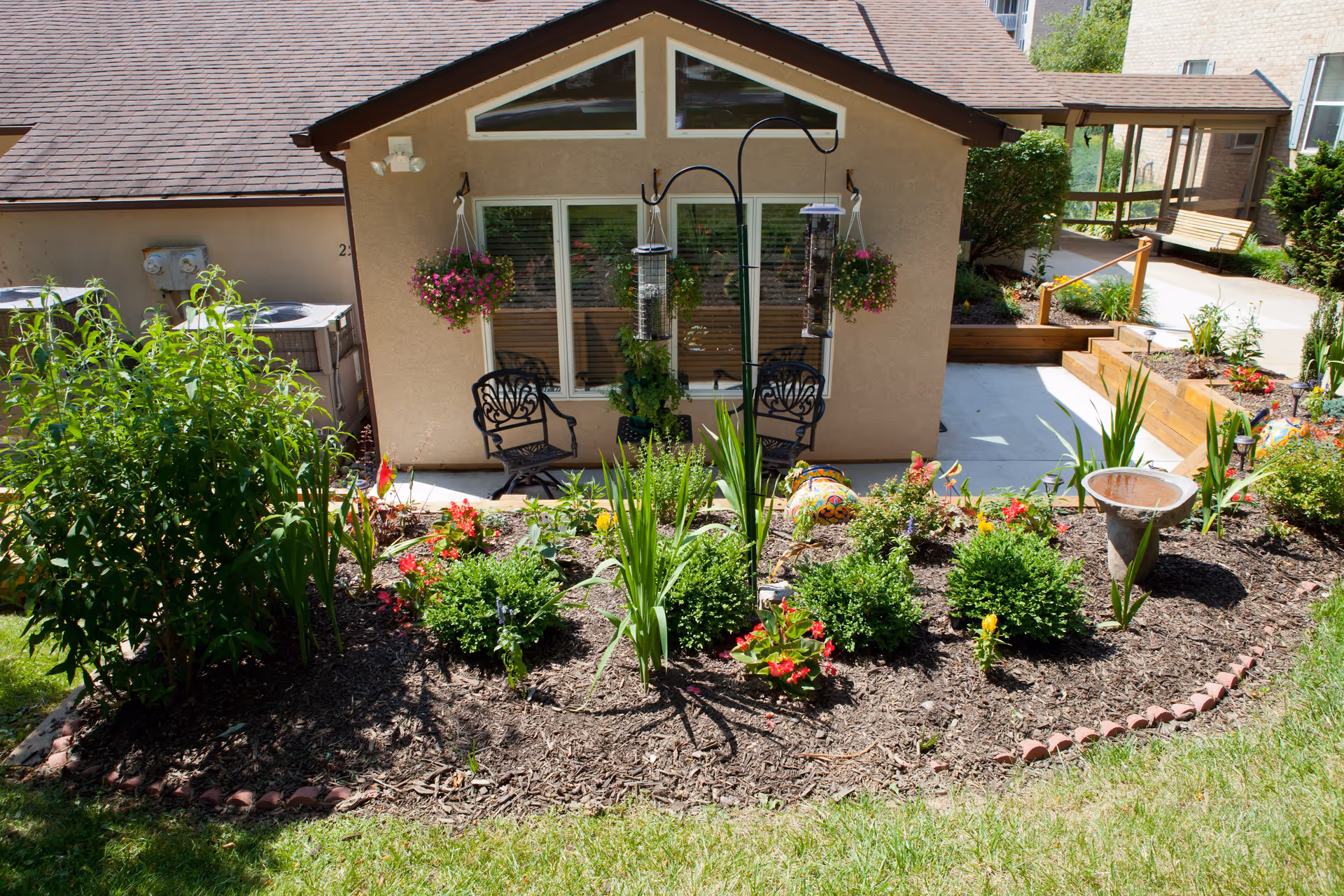 A small garden area with various green plants and colorful flowers in front of a beige building with a brown roof. Two black metal chairs are placed on a concrete patio near a window with white frames. Hanging flower pots with pink flowers are suspended on either side of the window. A bird feeder and a birdbath are also visible in the garden. The surrounding area includes a walkway and additional greenery.