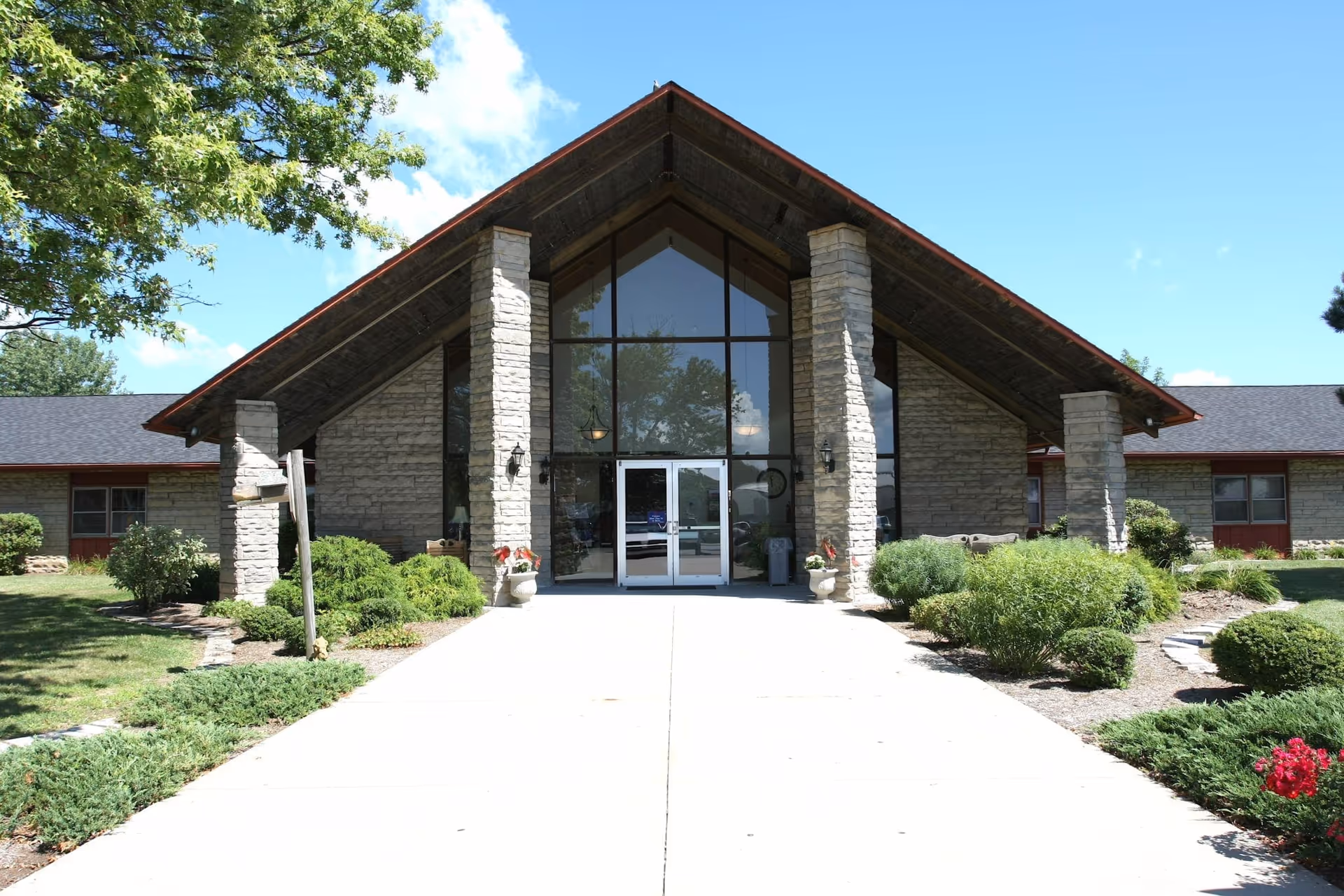 Front exterior view of Westview Nursing and Rehabilitation Center featuring a large peaked roof supported by stone pillars, glass entrance doors, and surrounding landscaped greenery under a clear blue sky.
