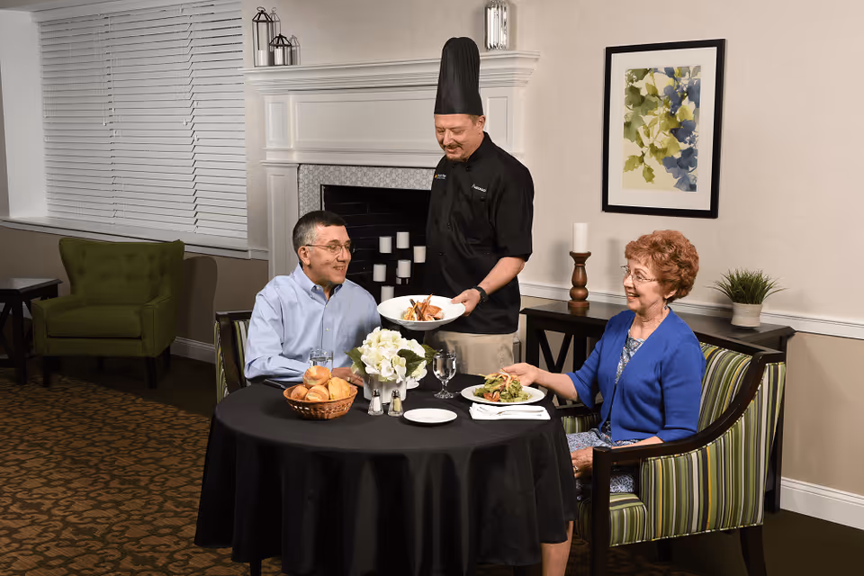 A chef in a black uniform and tall hat serves a plate of food to an elderly man and woman seated at a round table covered with a black tablecloth. The table has a basket of bread rolls, two glasses of water, and a floral centerpiece. The room features a fireplace with candles, a green armchair, a framed abstract painting, and a side table with a candle and a small plant.