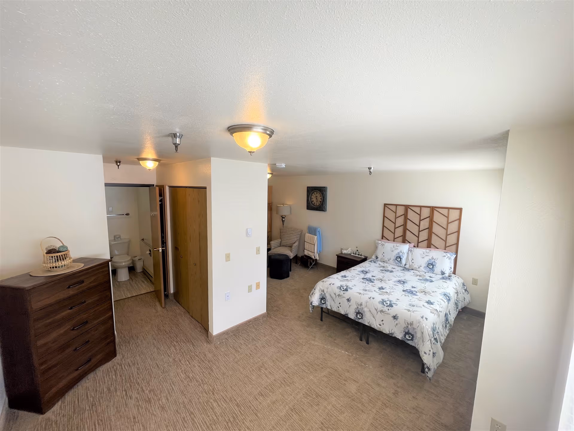 A senior living facility bedroom with a bed featuring floral bedding, a wooden headboard, a nightstand with a lamp, a cushioned chair with a small ottoman, a wall clock, a dresser with a basket on top, and an open door leading to a bathroom with a visible toilet.