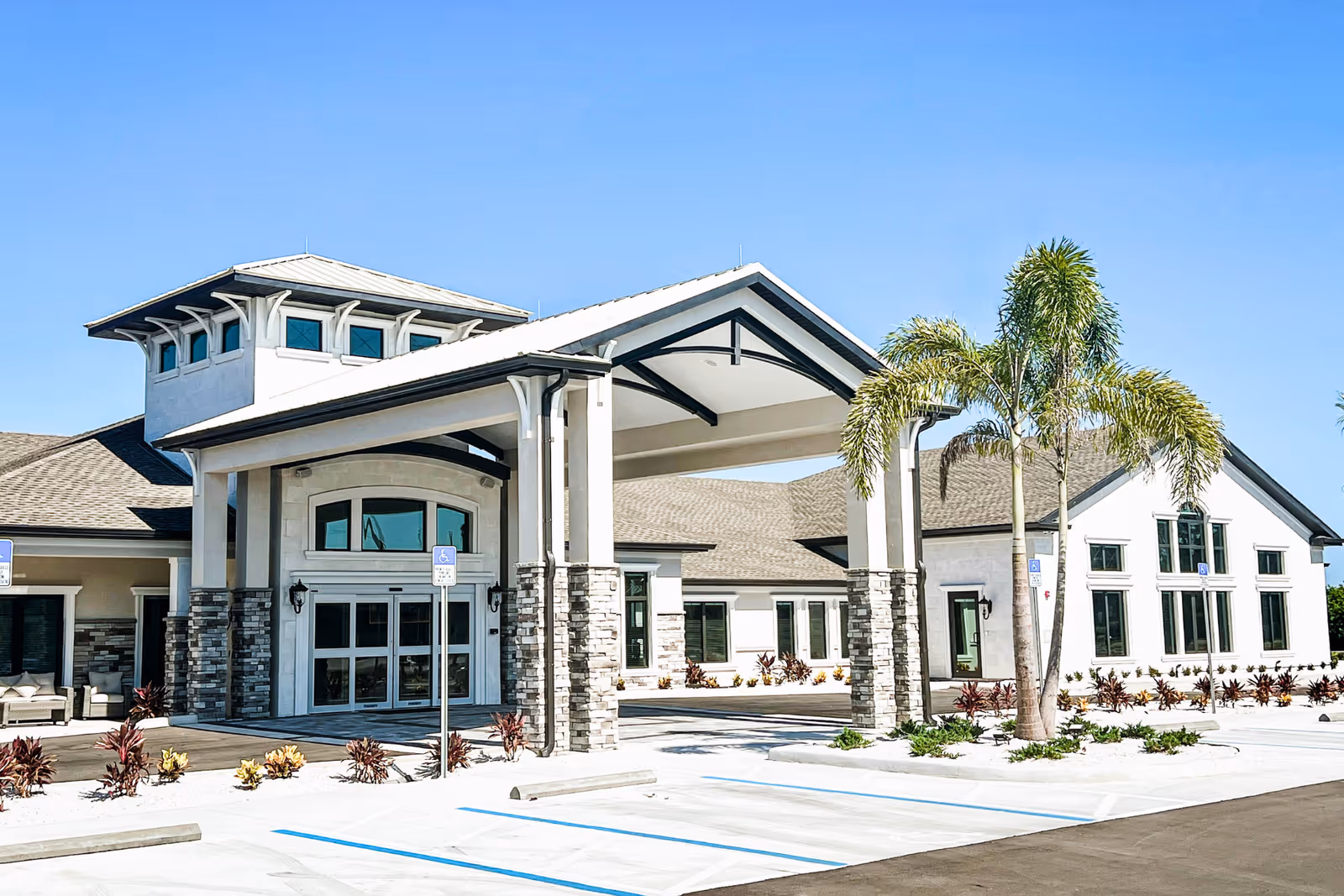 Front entrance of a light-colored senior living facility with a covered porte-cochere, palm trees, and marked parking spaces.