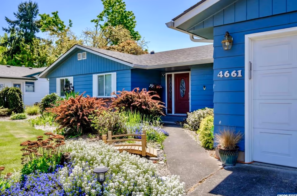 Front exterior of a single-story blue house with a landscaped garden, walkway, and a red front door showing house number 4661.