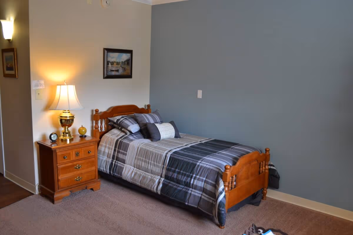 A cozy bedroom with a single wooden bed featuring a plaid comforter and multiple pillows. Next to the bed is a wooden nightstand with three drawers, a brass lamp with a white lampshade, a small clock, and a decorative item. The walls are painted in two tones, light beige and blue, with framed artwork hanging above the bed. The floor is carpeted.