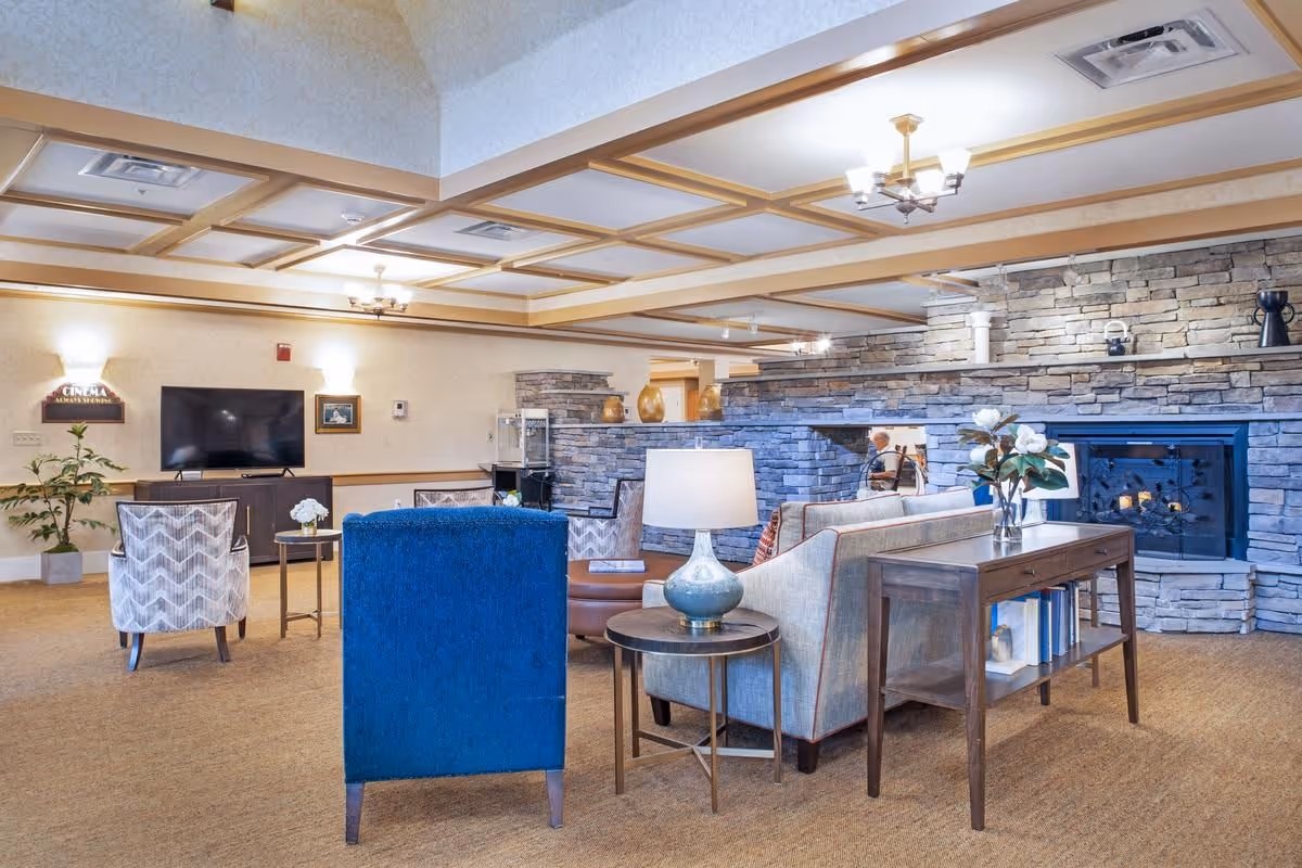 A cozy senior living facility common area with a stone fireplace, a TV mounted on the wall, comfortable seating including a blue armchair and patterned chairs, a wooden console table with books and a vase of flowers, and warm lighting from ceiling fixtures.