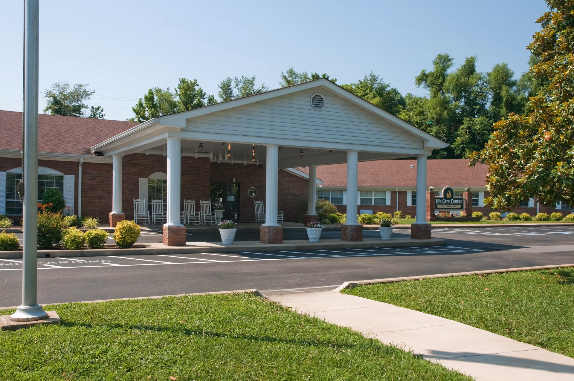 Front exterior view of Life Care Center of Centerville showing a covered entrance with white columns and rocking chairs, surrounded by a parking lot, green lawn, and trees under a clear blue sky.