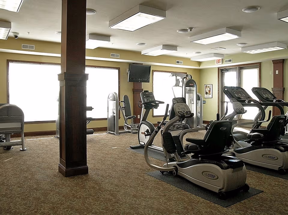 A fitness room with various exercise equipment including recumbent bikes, treadmills, and weight machines. The room has large windows letting in natural light, beige walls, carpeted floor, and ceiling lights. A TV is mounted on the wall near the windows.