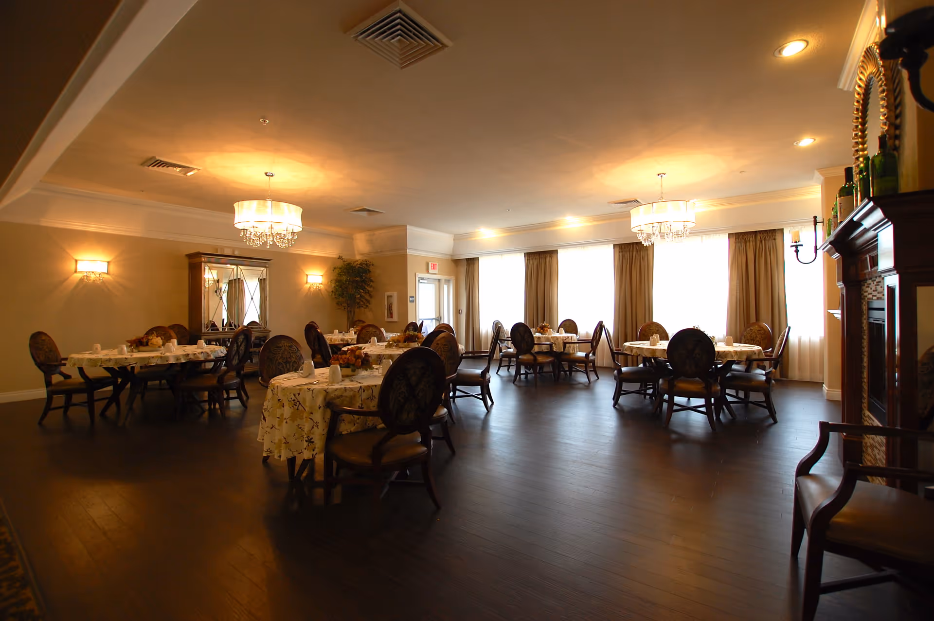 A warmly lit dining room with several round tables covered with floral tablecloths and set with cups and napkins. The room features dark wooden floors, upholstered chairs, chandeliers, wall sconces, large windows with curtains, and a decorative cabinet and fireplace.
