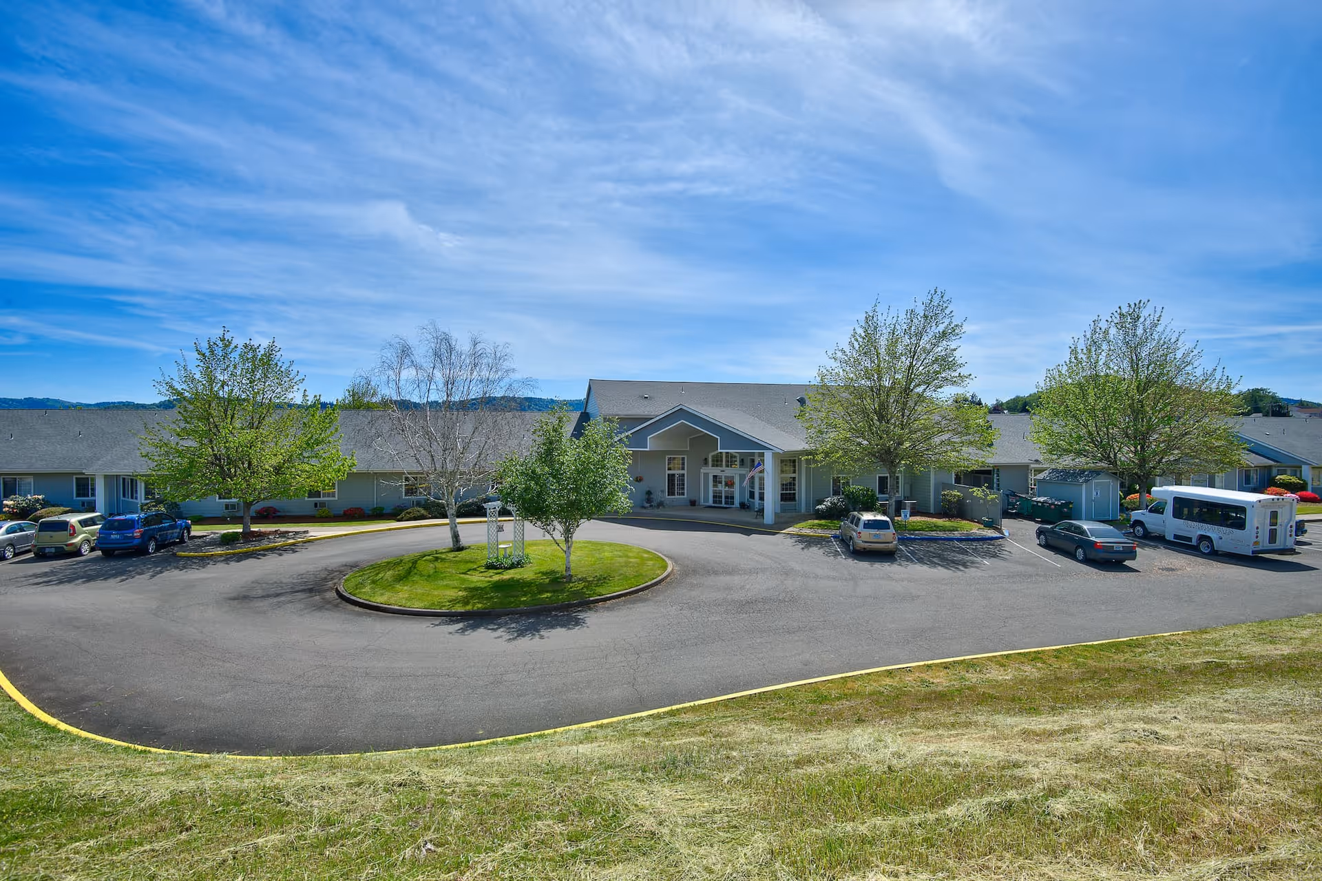 Front exterior view of Bridgewood Rivers Assisted Living facility with a circular driveway, several parked cars, trees, and a clear blue sky.