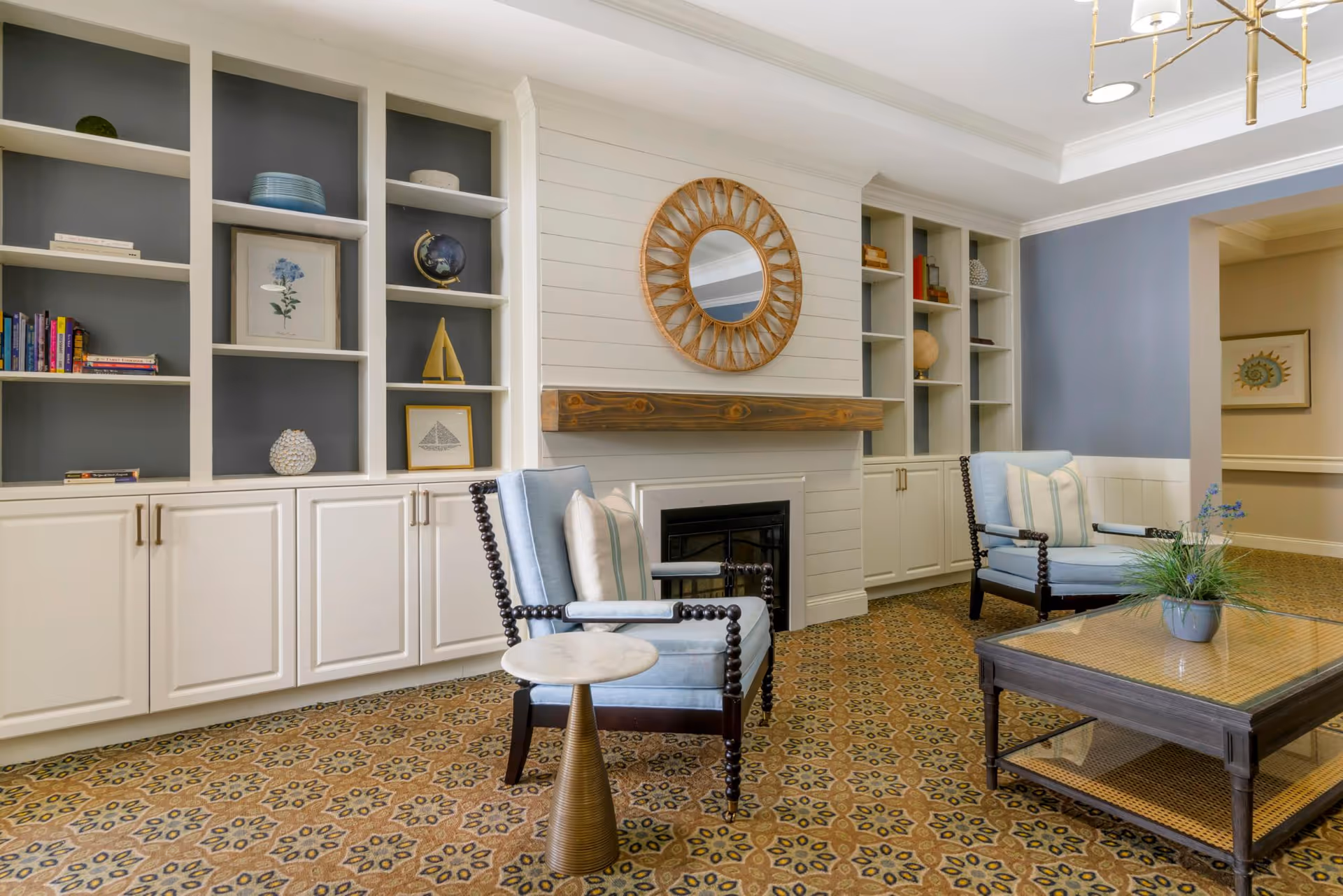 A cozy living room area featuring two light blue armchairs with white and blue striped pillows, a wooden coffee table with a plant on top, built-in white shelves with decorative items and books, a fireplace with a wooden mantel, and a round mirror above it. The room has patterned carpet flooring and soft blue and white walls.