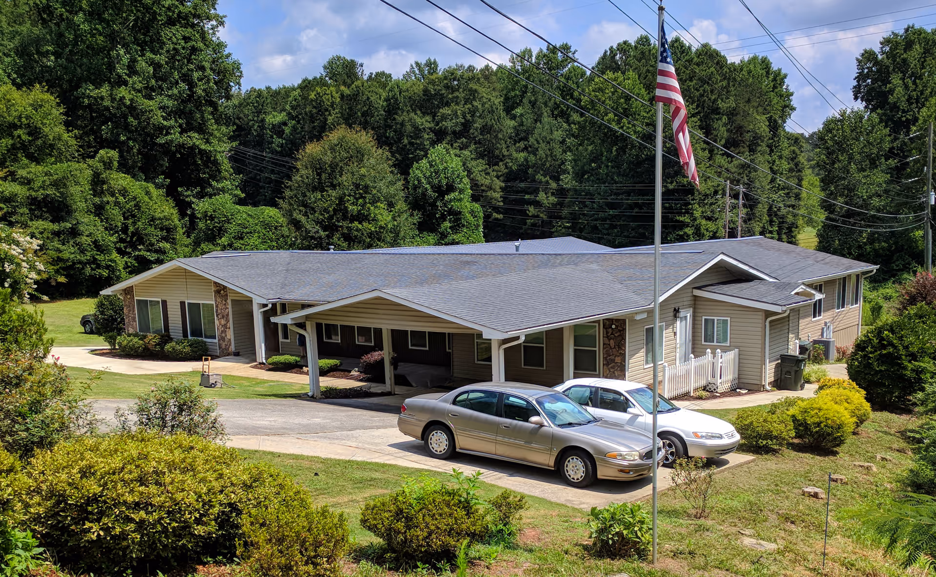 Exterior view of a single-story residential building with a gray roof and beige siding, surrounded by green trees and bushes. Two cars are parked in front of the building near a flagpole with an American flag.