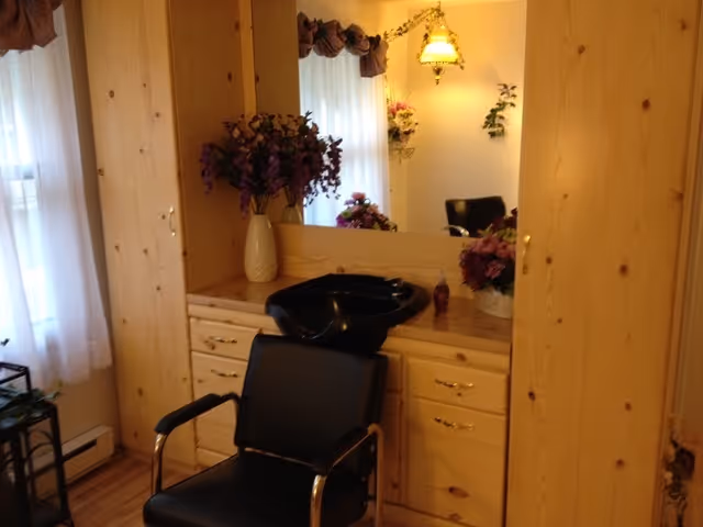 Interior salon corner with a black salon chair and hair-washing basin set against wooden cabinets and a mirror, decorated with flowers.