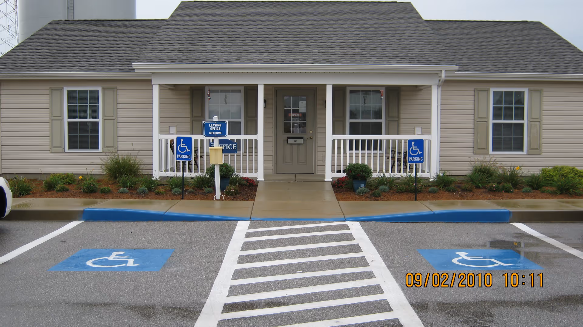 Exterior view of a single-story building with beige siding and a gray roof. The building has a small porch with white railings and a central door flanked by two windows. There are two handicap parking spaces in front of the building, marked with blue paint and signs. A sign near the door reads 'Leasing Office Welcome'.