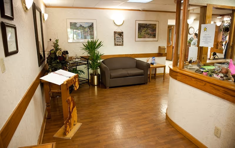 A waiting area inside Bethlehem Woods Nursing and Rehabilitation featuring a brown loveseat, a small side table with a box of tissues, a plant in a gold pot, framed pictures on the wall, a wooden stand with a guest book, and a reception window with various items and a person visible behind it.