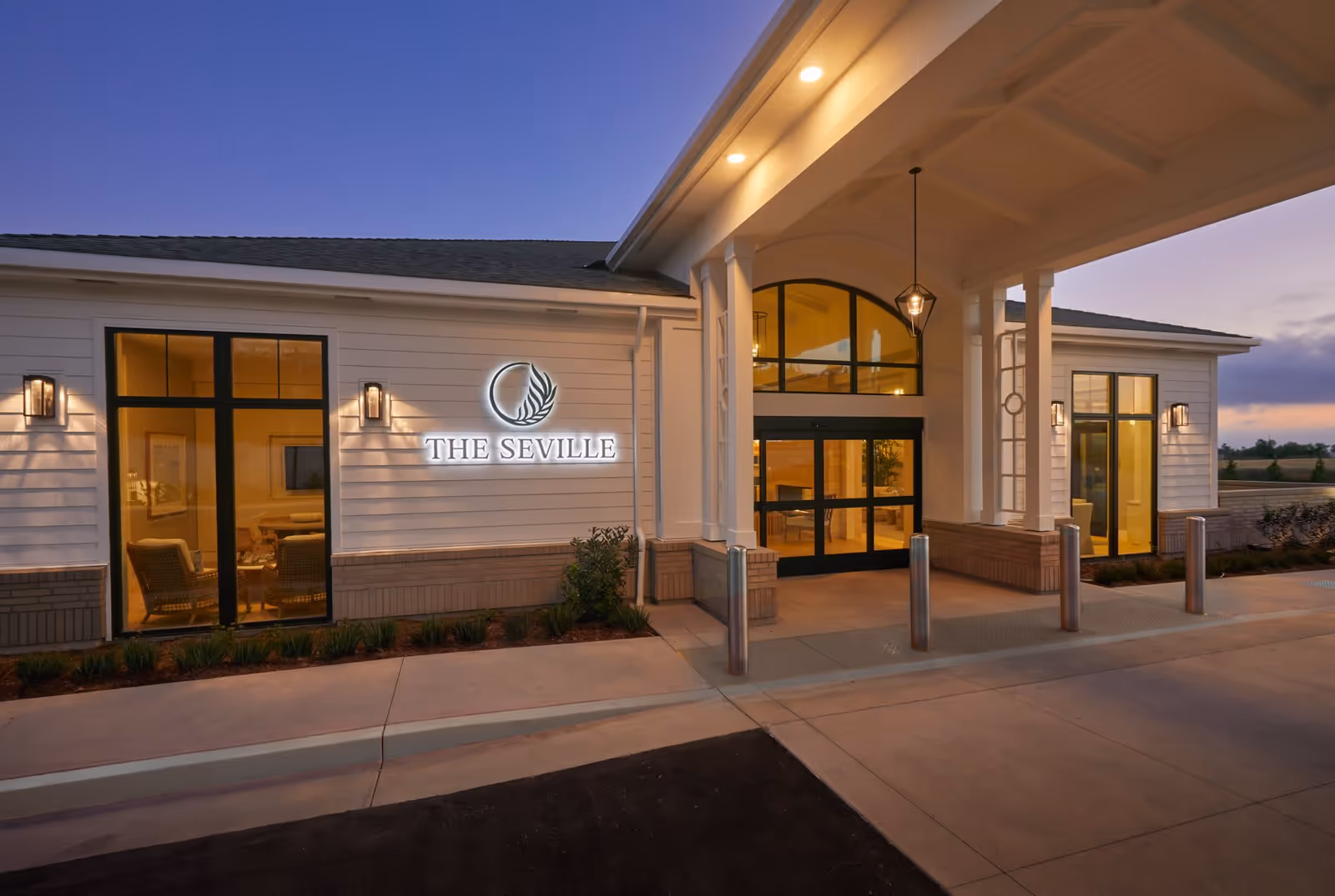 Exterior view of The Seville senior living facility at dusk, showing the entrance with large windows, illuminated signage, and a covered walkway with pillars and lights.