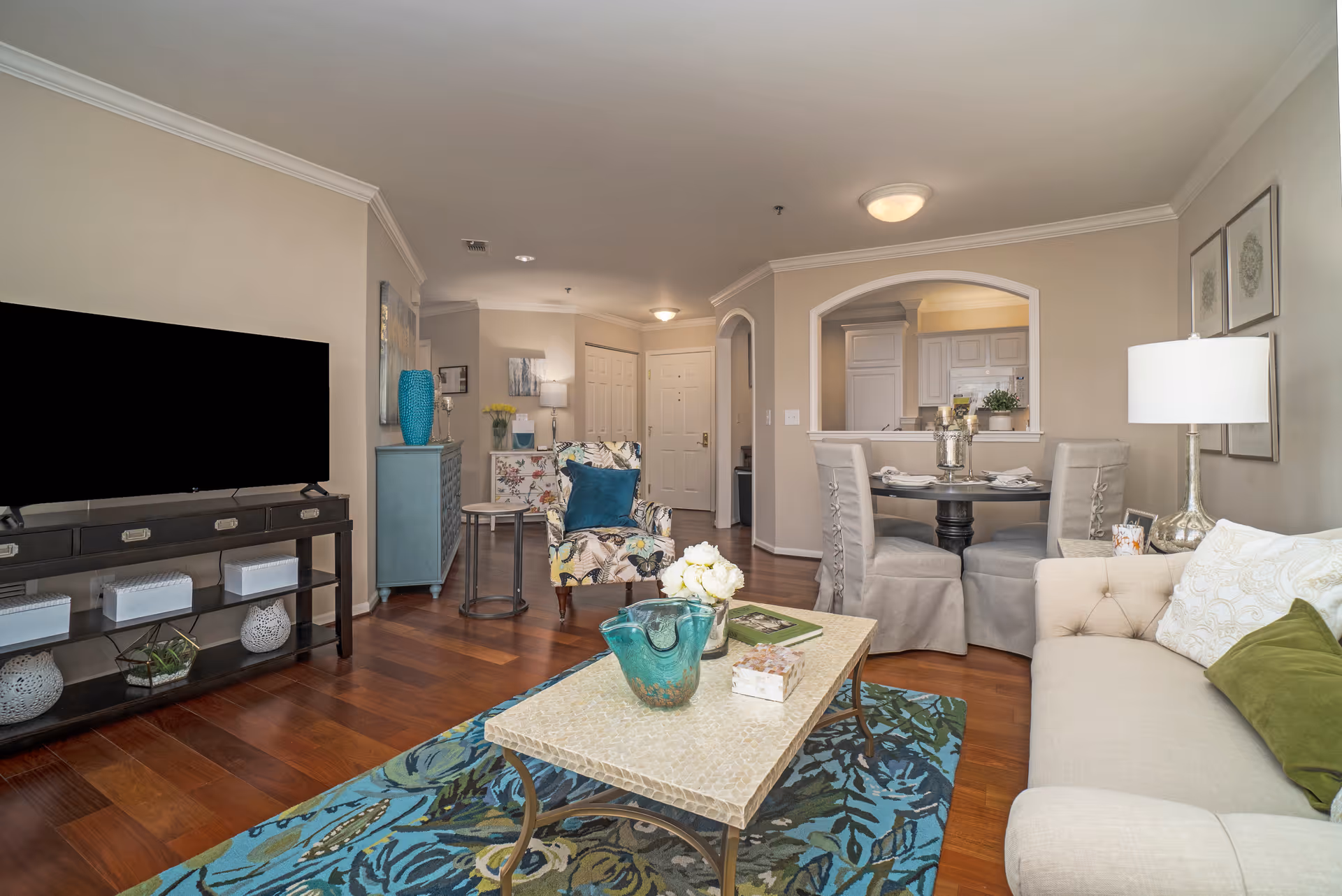 Bright living room interior featuring a sofa, coffee table, TV console, patterned rug, and a dining nook with an arched pass-through to the kitchen.