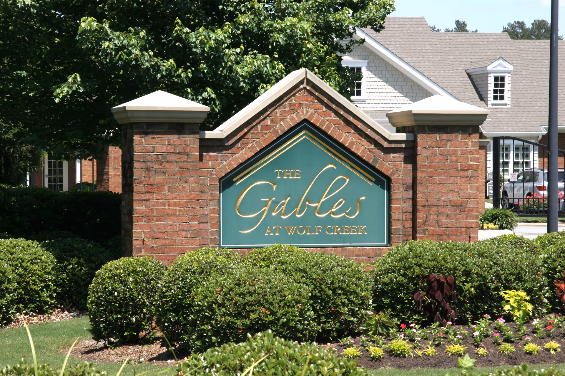 A brick sign with a green and gold plaque that reads 'The Gables at Wolf Creek' surrounded by neatly trimmed bushes and landscaping, with a building and trees in the background.