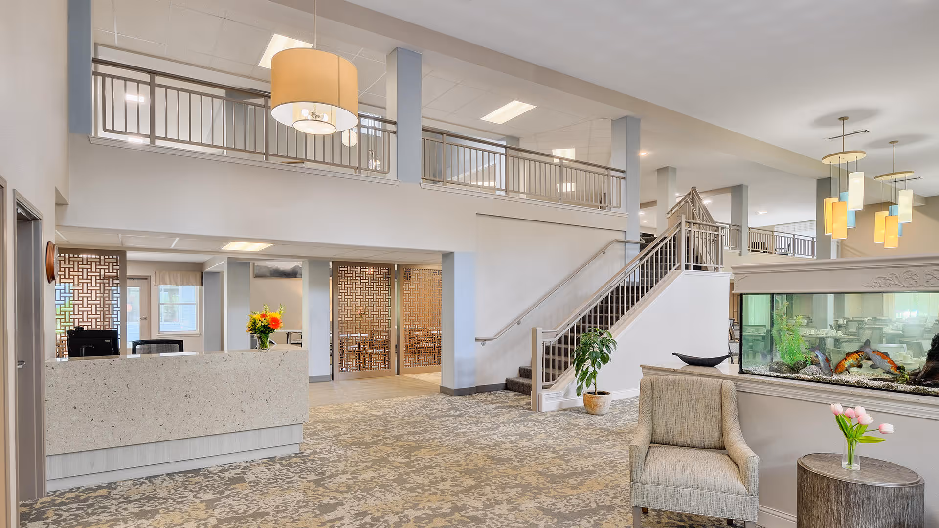 Spacious and well-lit senior living facility lobby with a reception desk on the left, decorative wooden partitions, a staircase leading to an upper level, a large fish tank on the right, and comfortable seating with a small table holding a vase of pink tulips.