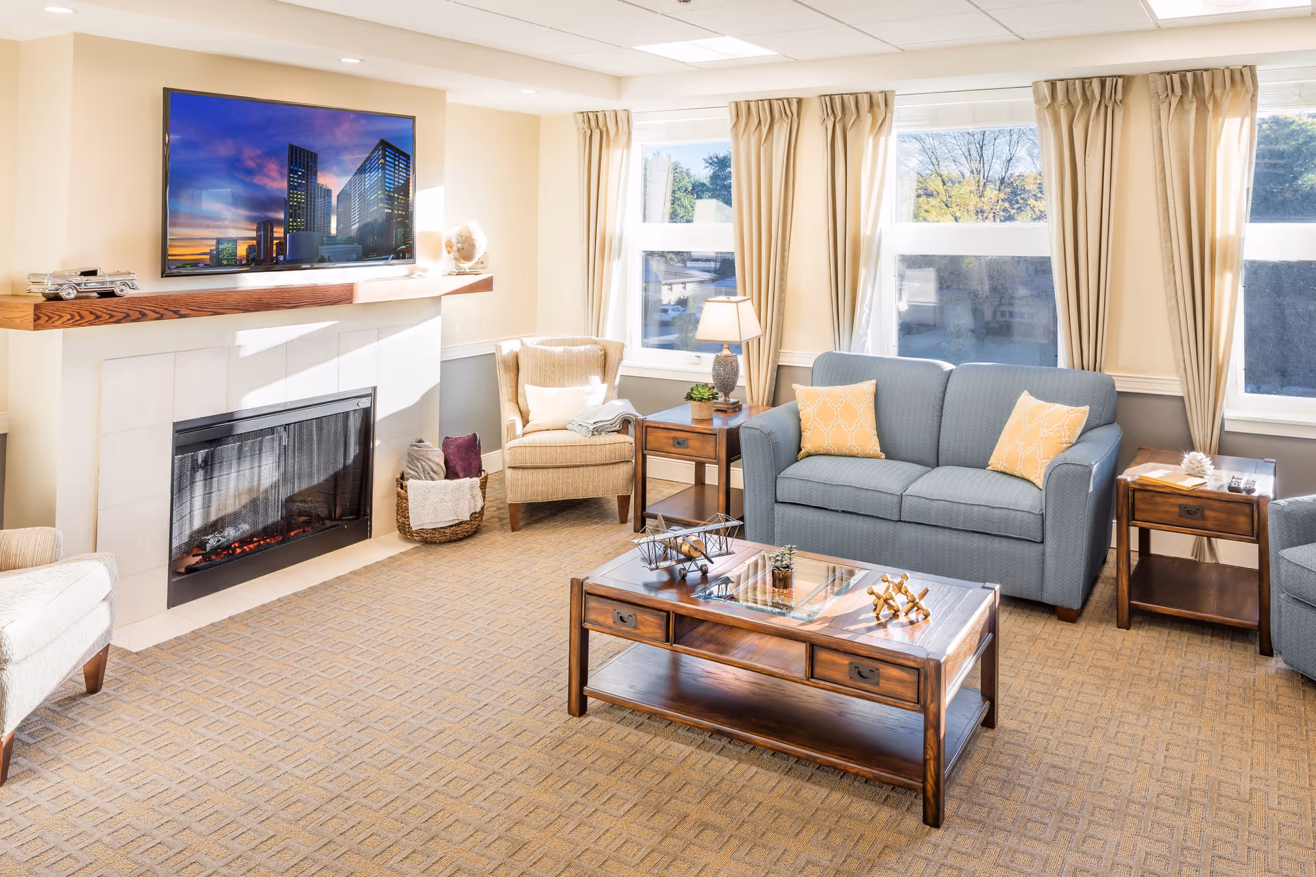 A bright and cozy living room with a blue sofa adorned with two yellow patterned pillows, two beige armchairs, a wooden coffee table with decorative items, and two wooden side tables with a lamp and books. The room features large windows with beige curtains allowing natural light to fill the space, and a fireplace with a mounted flat-screen TV above it displaying a cityscape at sunset.