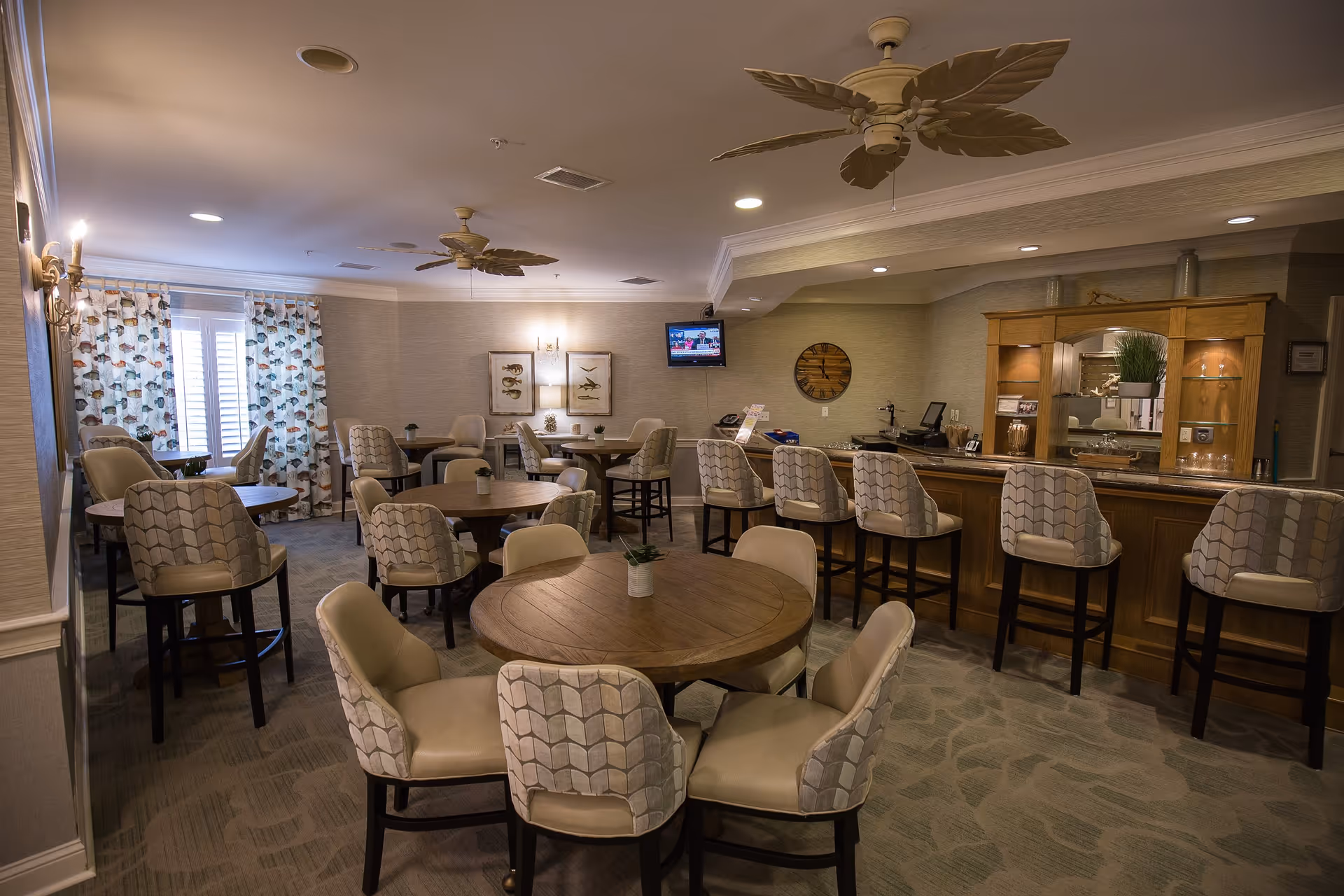 A cozy dining area in a senior living facility with several round wooden tables surrounded by cushioned chairs. There is a bar counter with high chairs on the right side, a wall-mounted TV, decorative wall art, and ceiling fans with leaf-shaped blades. The room has soft lighting, patterned curtains, and a carpeted floor.