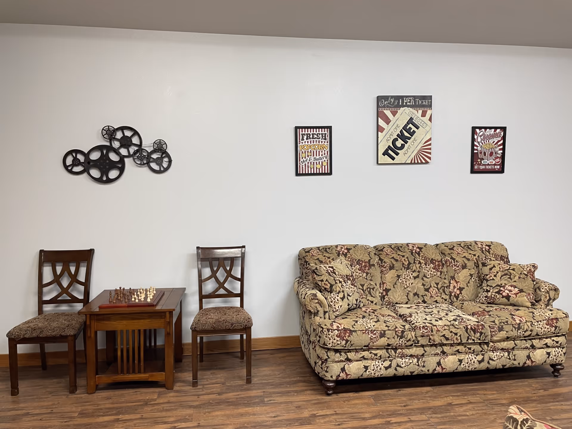 A cozy room with a floral patterned sofa on the right and a small wooden table with a chessboard between two wooden chairs on the left. The wall behind has decorative movie-themed wall art including film reels and vintage-style posters.