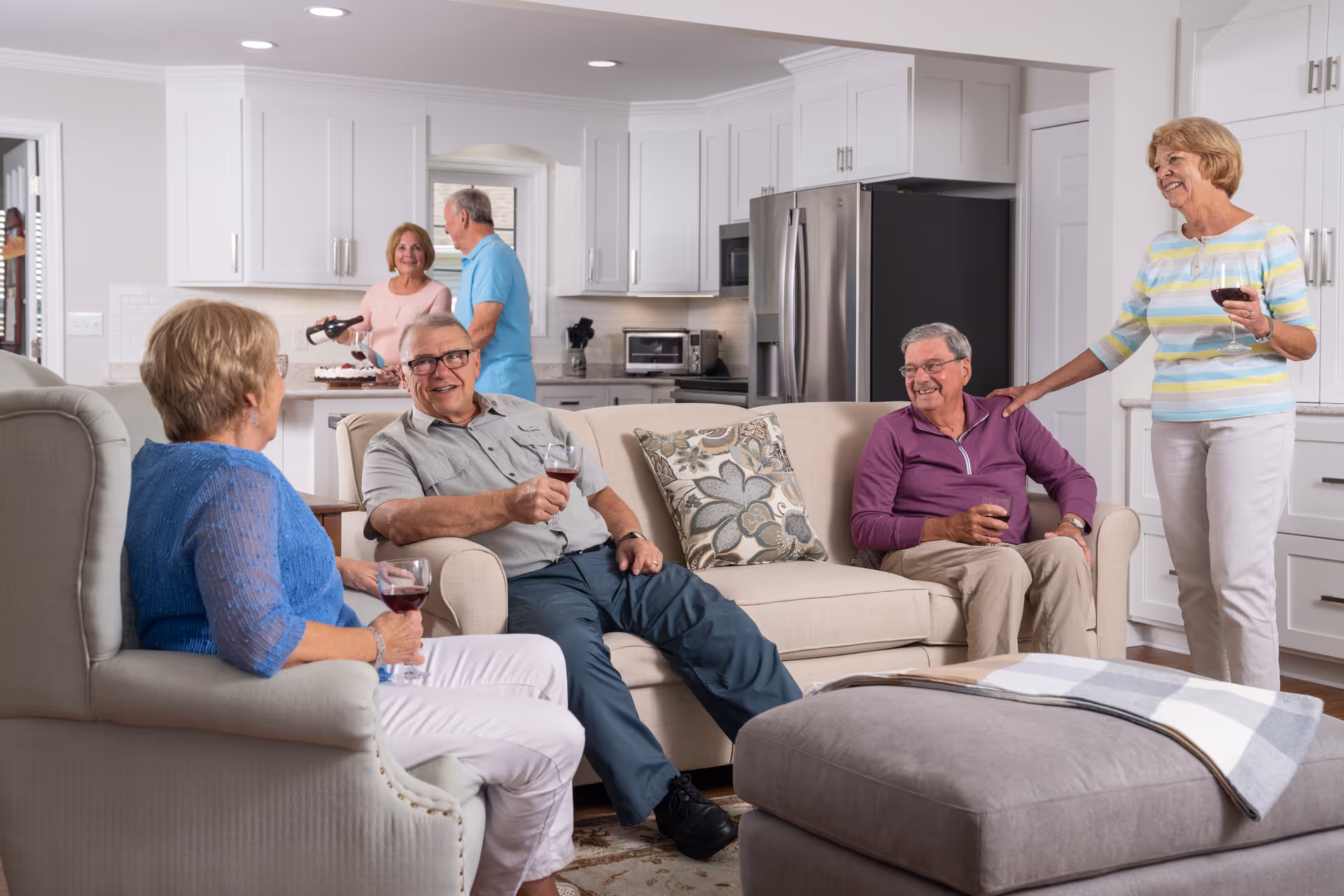 A group of five elderly people socializing in a modern, bright kitchen and living room area. Three people are seated on sofas holding glasses of red wine, while two others stand in the kitchen area, one pouring wine. The room features white cabinets, stainless steel refrigerator, and a cozy, inviting atmosphere.