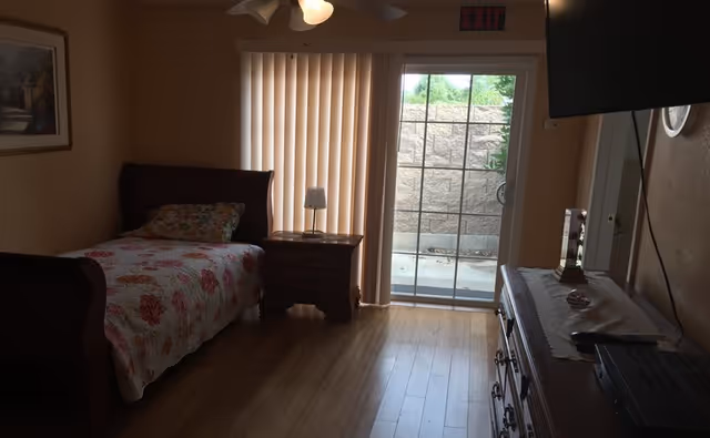 A small bedroom with a single bed covered in a floral quilt, a wooden nightstand with a lamp, vertical blinds covering a sliding glass door leading outside, a wooden dresser with a TV mounted above it, and a ceiling fan with lights.