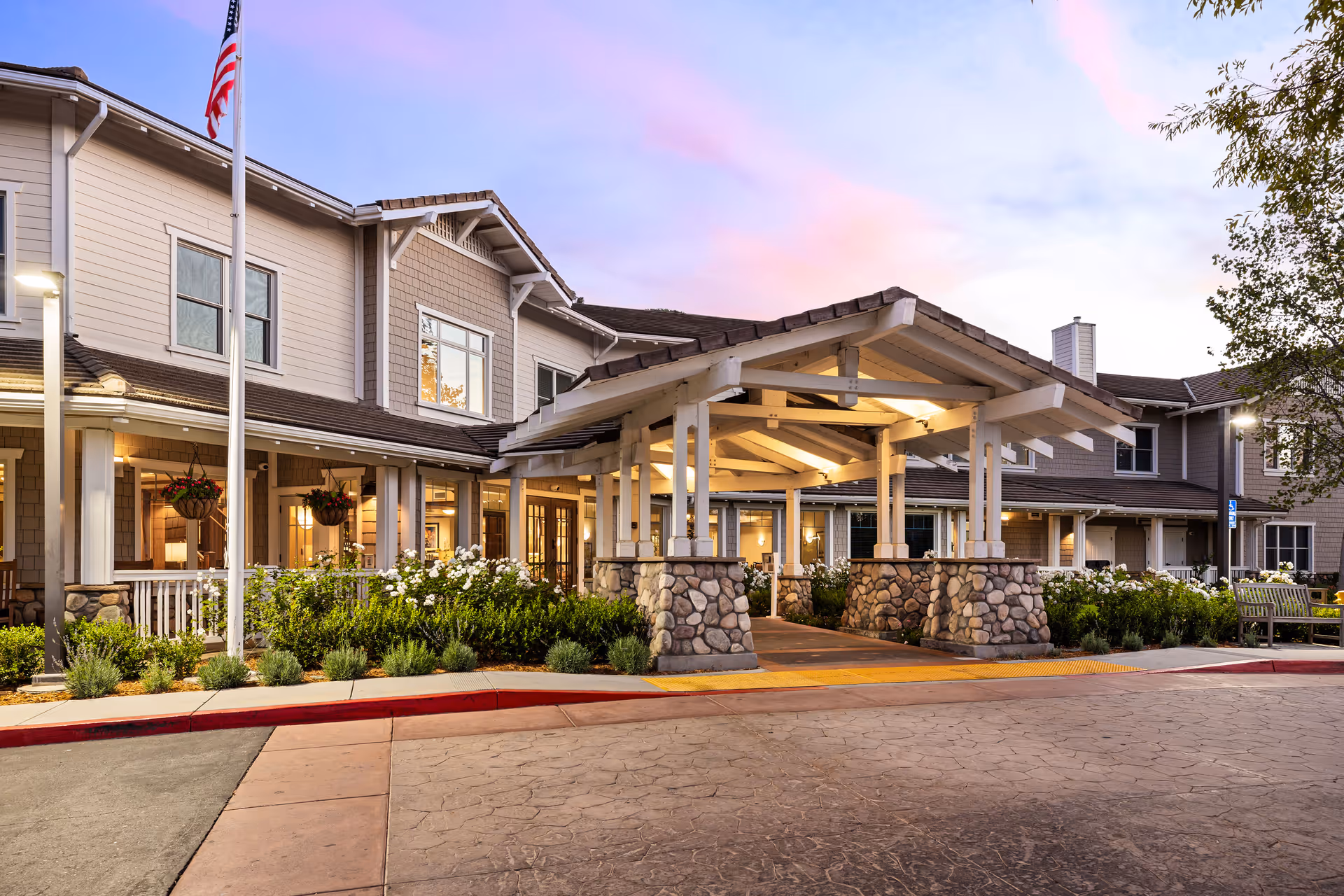 Exterior view of a senior living facility building at dusk with a covered entrance supported by stone pillars, well-maintained landscaping with bushes and flowers, an American flag on a flagpole, and warm interior lights visible through the windows.