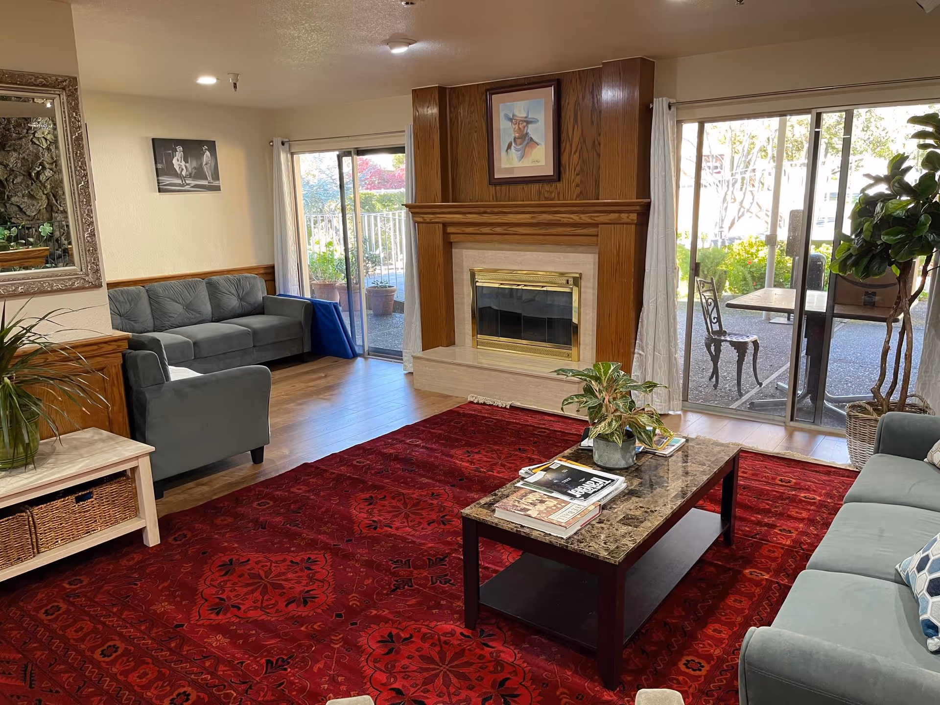 A cozy living room with a red patterned rug covering the wooden floor. There are two gray sofas and a matching armchair arranged around a rectangular coffee table with a marble top. The coffee table holds some magazines and a potted plant. A wooden fireplace with a brass frame is centered on the far wall, above which hangs a portrait of a man wearing a hat. Large sliding glass doors on either side of the fireplace let in natural light and provide views of an outdoor patio area with chairs and plants. A large mirror and a black and white photograph hang on the walls, and there are additional plants placed around the room.