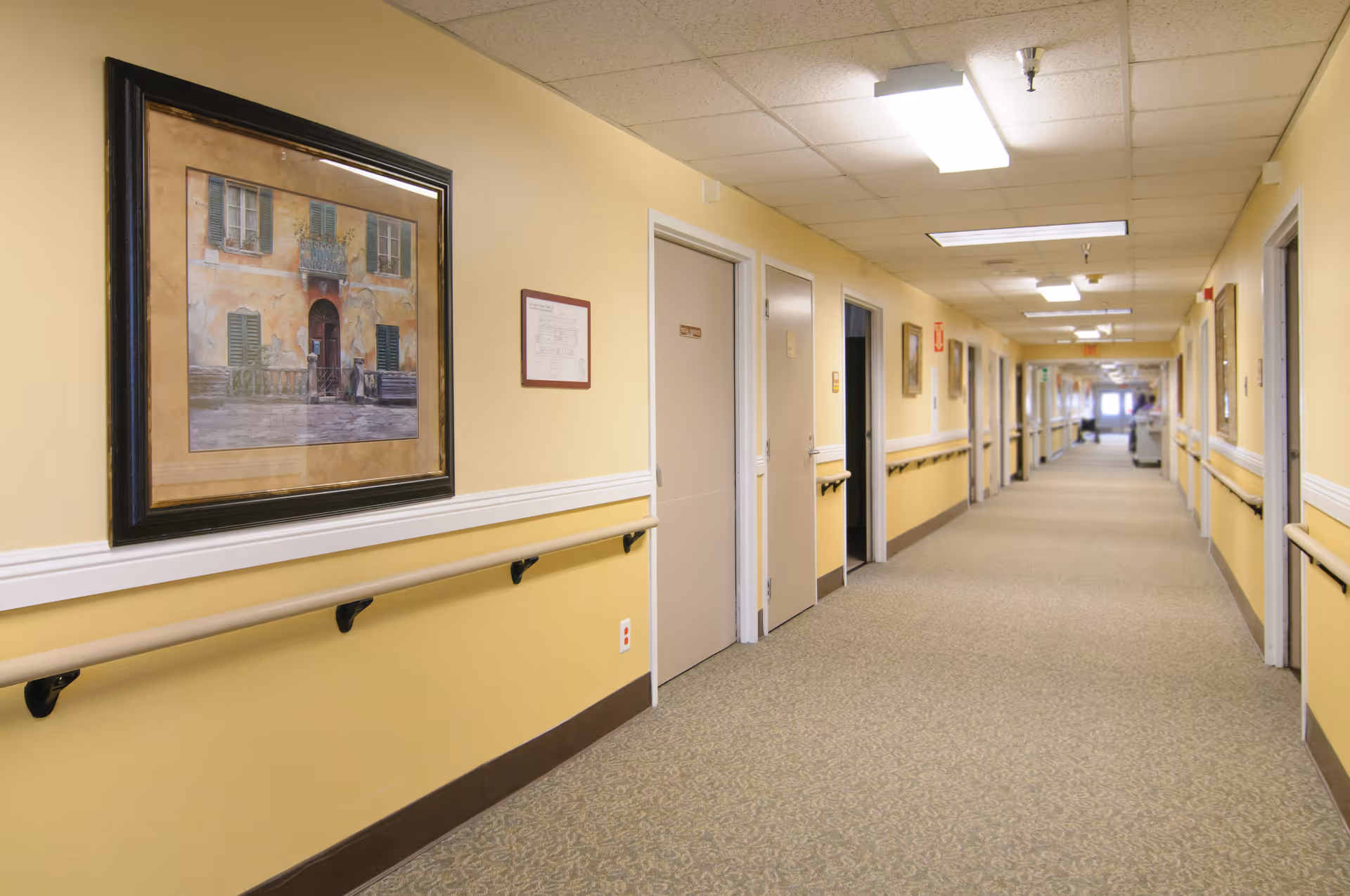 Long, well-lit interior hallway of a senior living facility with handrails, framed artwork, and multiple doors along the corridor.