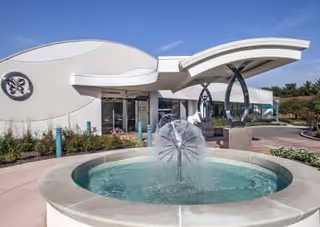 Entrance of a senior living facility featuring a round fountain in front of a curved canopy and modern building facade.