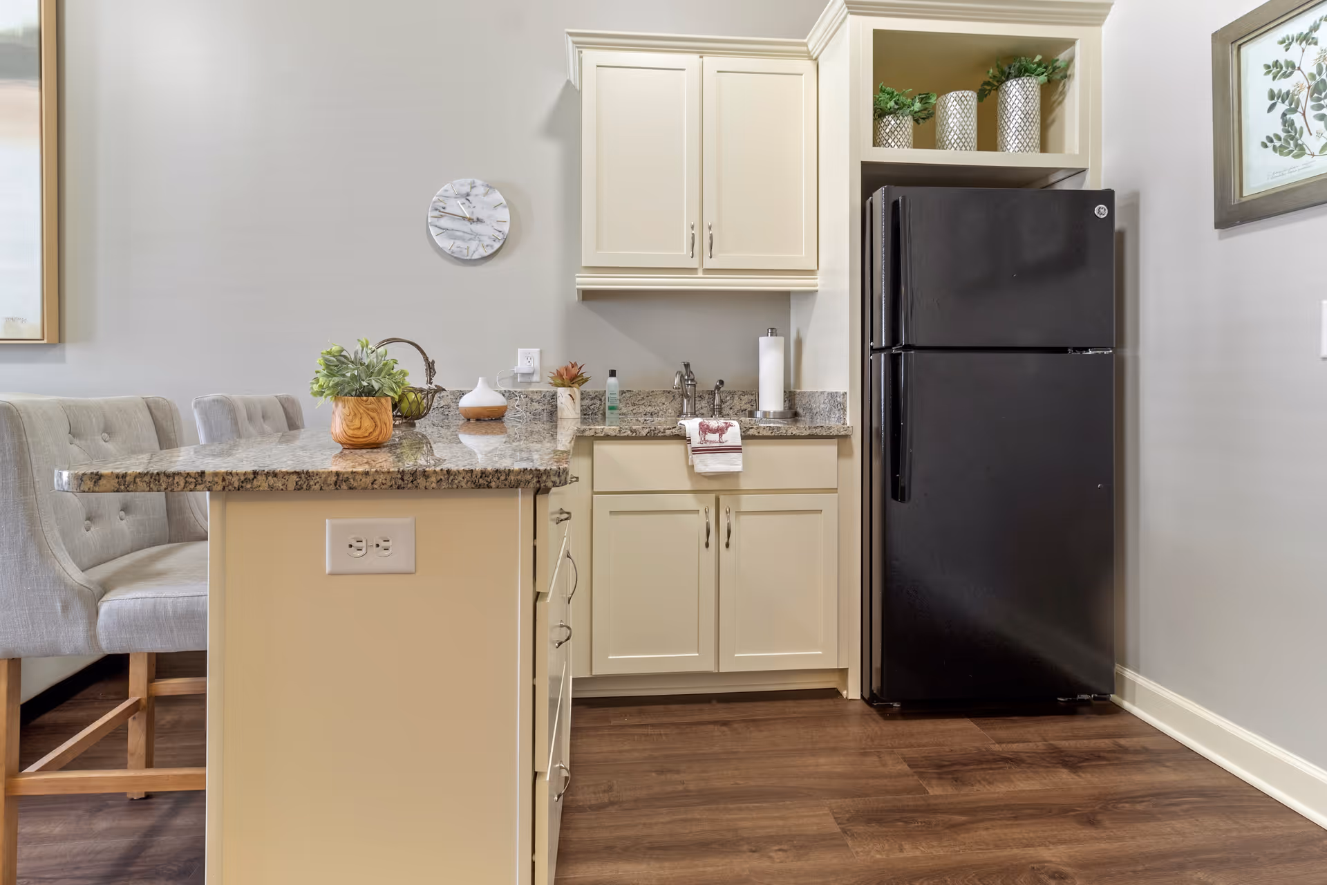 A modern kitchen area with cream-colored cabinets, a black refrigerator, and a granite countertop island with two upholstered chairs. The countertop has decorative plants and a diffuser. A wall clock and framed artwork are visible on the walls.