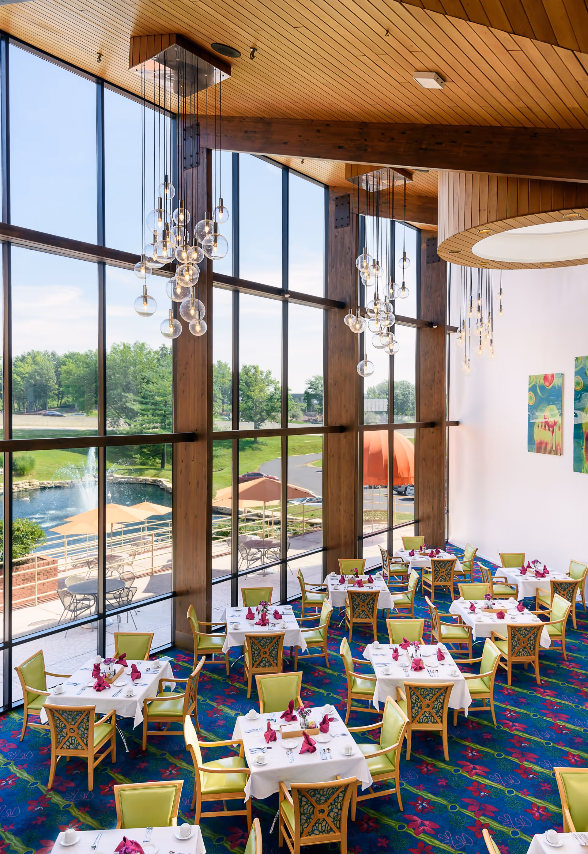 A bright dining room with large floor-to-ceiling windows offering a view of a pond, trees, and outdoor seating area. The room features multiple tables covered with white tablecloths, each set with cups, plates, and folded red napkins. The chairs have green cushions and decorative backs. Modern hanging light fixtures with multiple glass bulbs hang from a wooden ceiling.