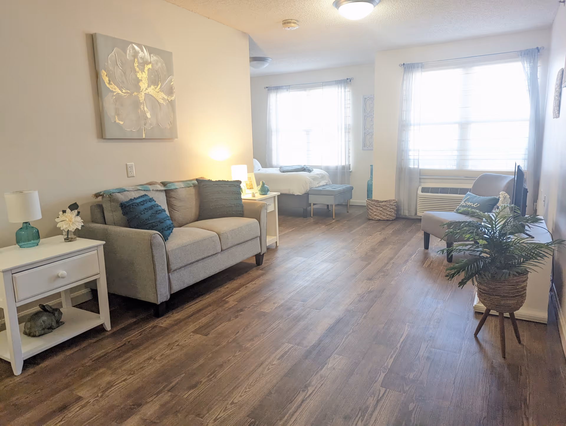 A bright and cozy living space featuring a gray sofa with blue and gray cushions, a white side table with a lamp and decorative items, a small armchair with a 'home' pillow, a TV on a stand, a potted plant, and a bed in the background near large windows with sheer curtains.
