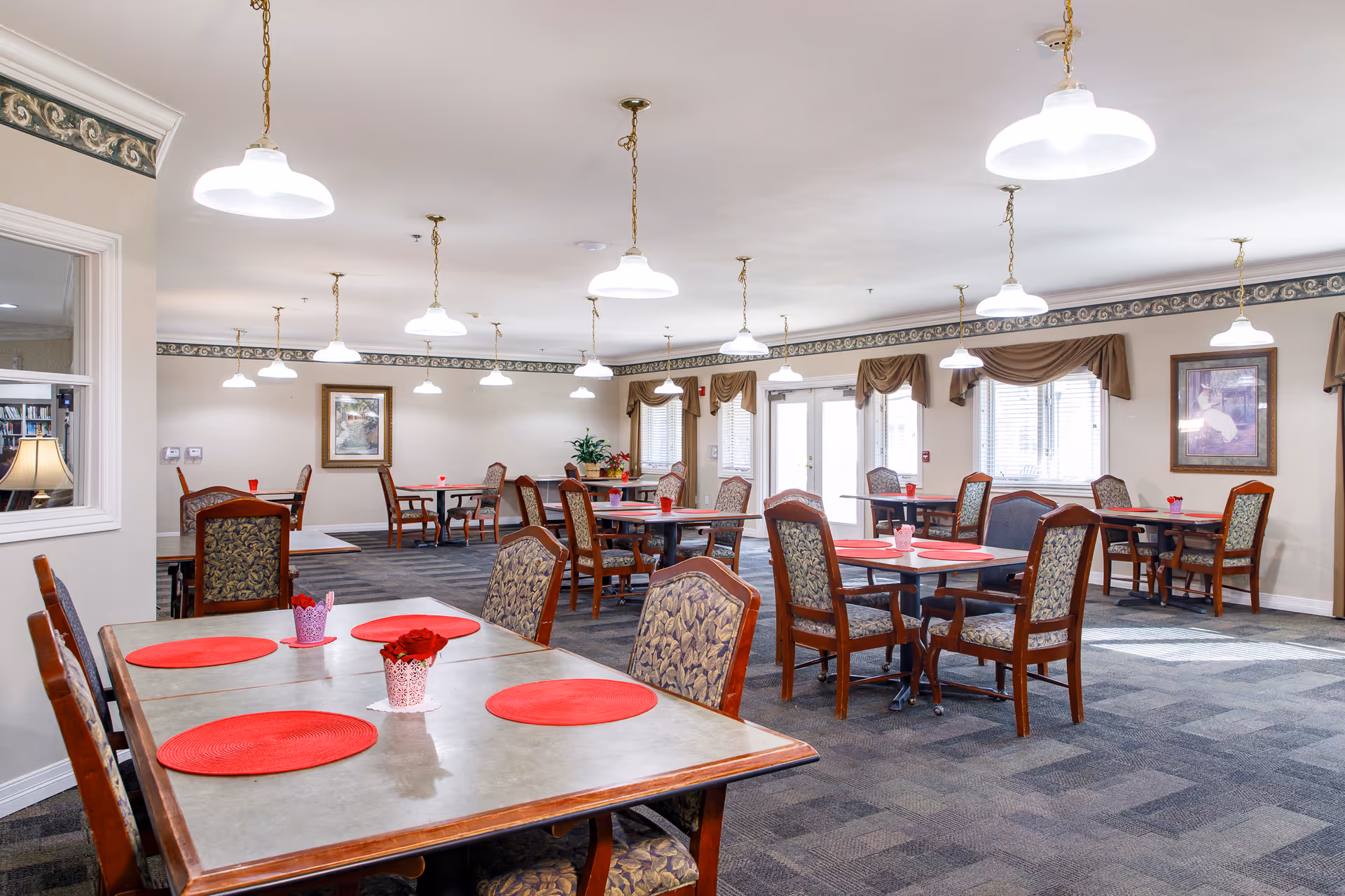 Bright communal dining room with several tables and upholstered chairs, red place mats, hanging pendant lights, and framed artwork.