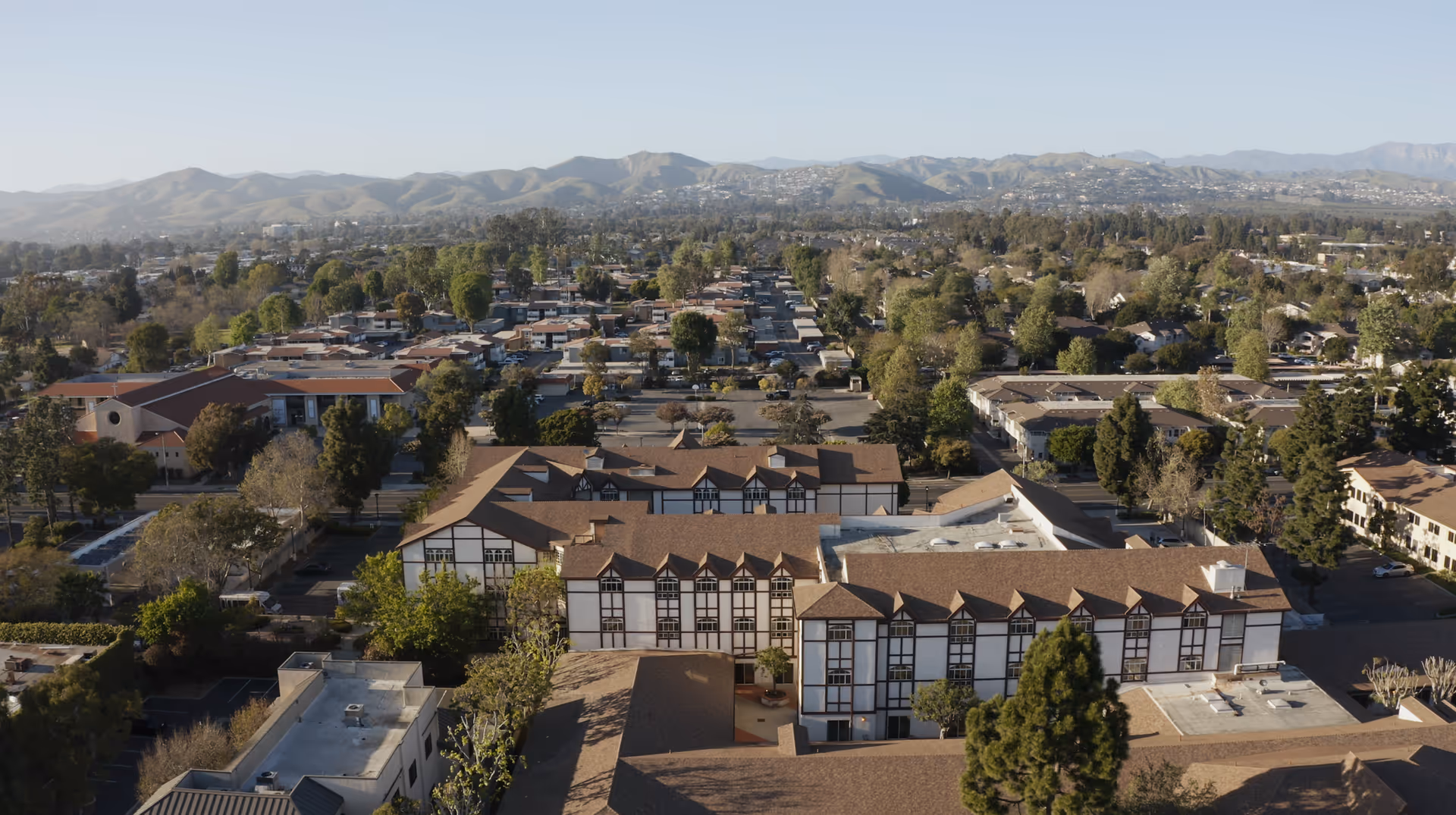 Aerial view of a large assisted living facility with multiple connected buildings featuring brown roofs and white walls with dark trim, surrounded by trees and residential neighborhoods, with mountains visible in the background under a clear sky.