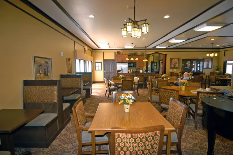 A spacious dining room in a senior living facility with multiple wooden tables and chairs arranged neatly. The tables have small flower arrangements as centerpieces. There are booth-style seating areas along the left wall and a kitchen area visible in the background. The room is warmly lit with ceiling lights and decorated with framed artwork on the walls.