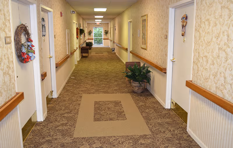 A long, carpeted hallway in a senior living facility with beige patterned wallpaper and white doors on both sides, some decorated with wreaths and hanging ornaments. There are handrails along the walls, potted plants, framed artwork, and a window at the far end letting in natural light.
