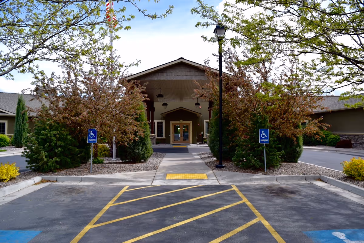 Entrance to Quinn Meadows Rehabilitation and Care Center with a covered walkway, surrounded by trees and shrubs. There are two handicap parking signs on either side of the walkway, and a flagpole with an American flag is visible behind the trees.