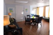 Well-lit common room with a dining table and chairs, sofas, lamps, framed art, and large windows with curtains.