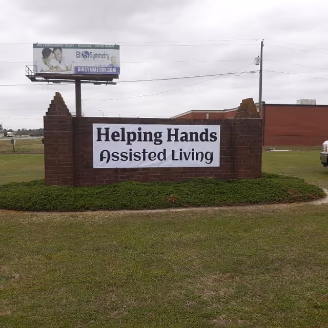 Brick sign on a lawn reading 'Helping Hands Assisted Living' with a building and billboard in the background.