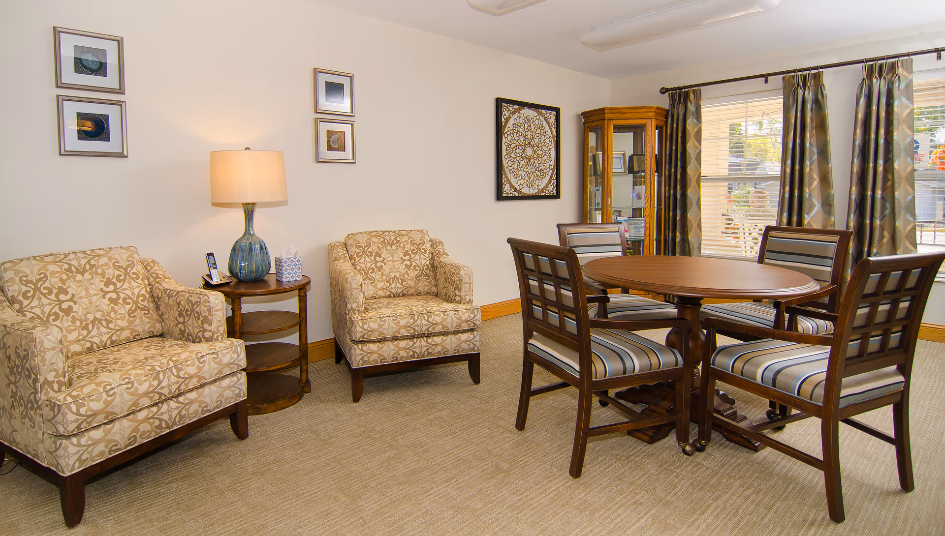 A cozy sitting area with two patterned armchairs and a wooden side table with a lamp and a phone. Adjacent to the sitting area is a round wooden table with four striped cushioned chairs. The room has beige walls adorned with framed artwork, a glass display cabinet, and large windows with patterned curtains letting in natural light.