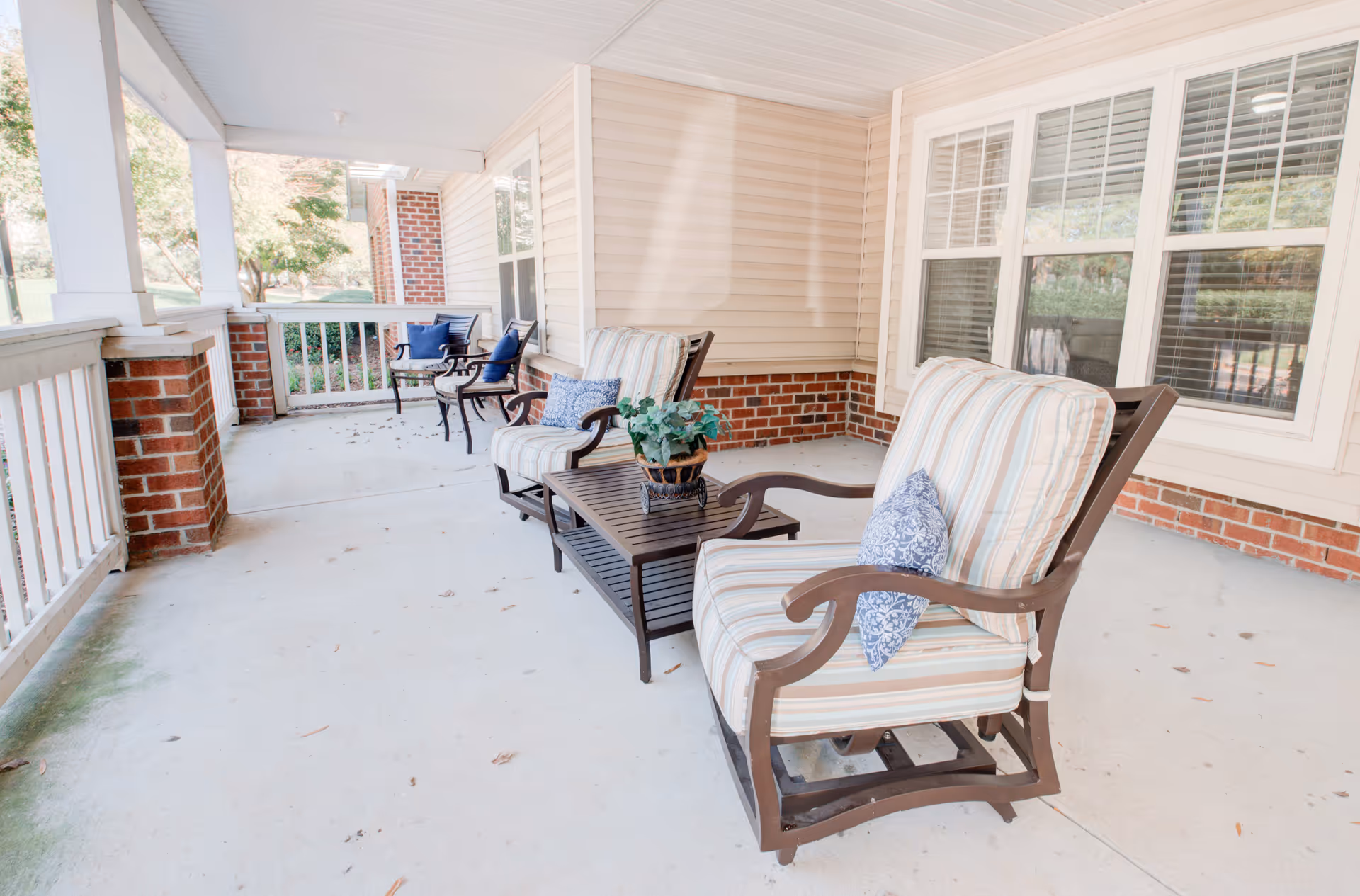 Covered outdoor patio area with cushioned armchairs and a coffee table with a potted plant. The patio has white railings, brick pillars, and beige siding walls with windows.
