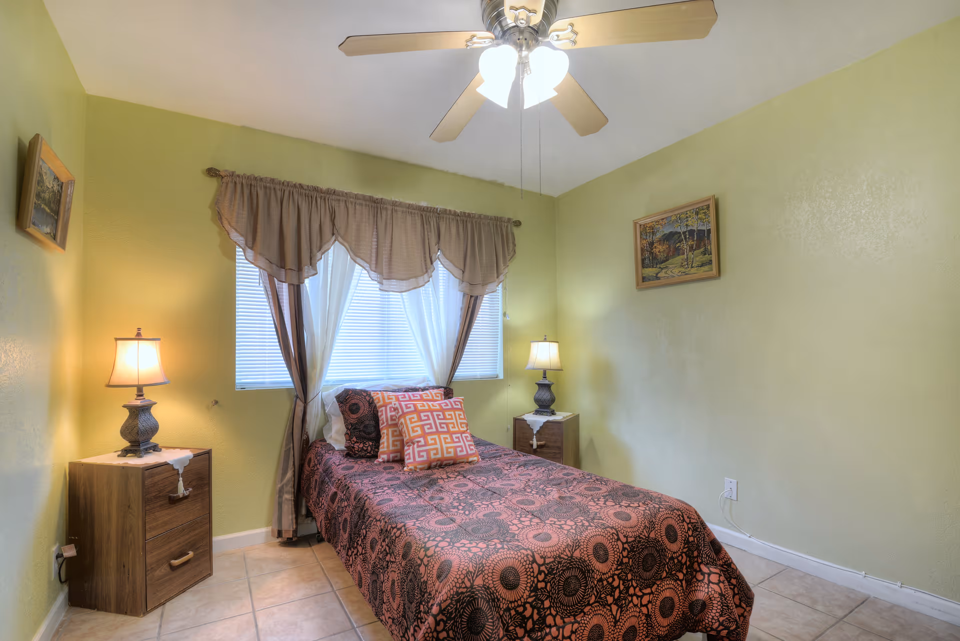 A small bedroom with light green walls and tiled floor. The room features a single bed with a patterned bedspread and decorative pillows, two wooden nightstands each with a lamp, a window with curtains, a ceiling fan with lights, and two framed paintings on the walls.