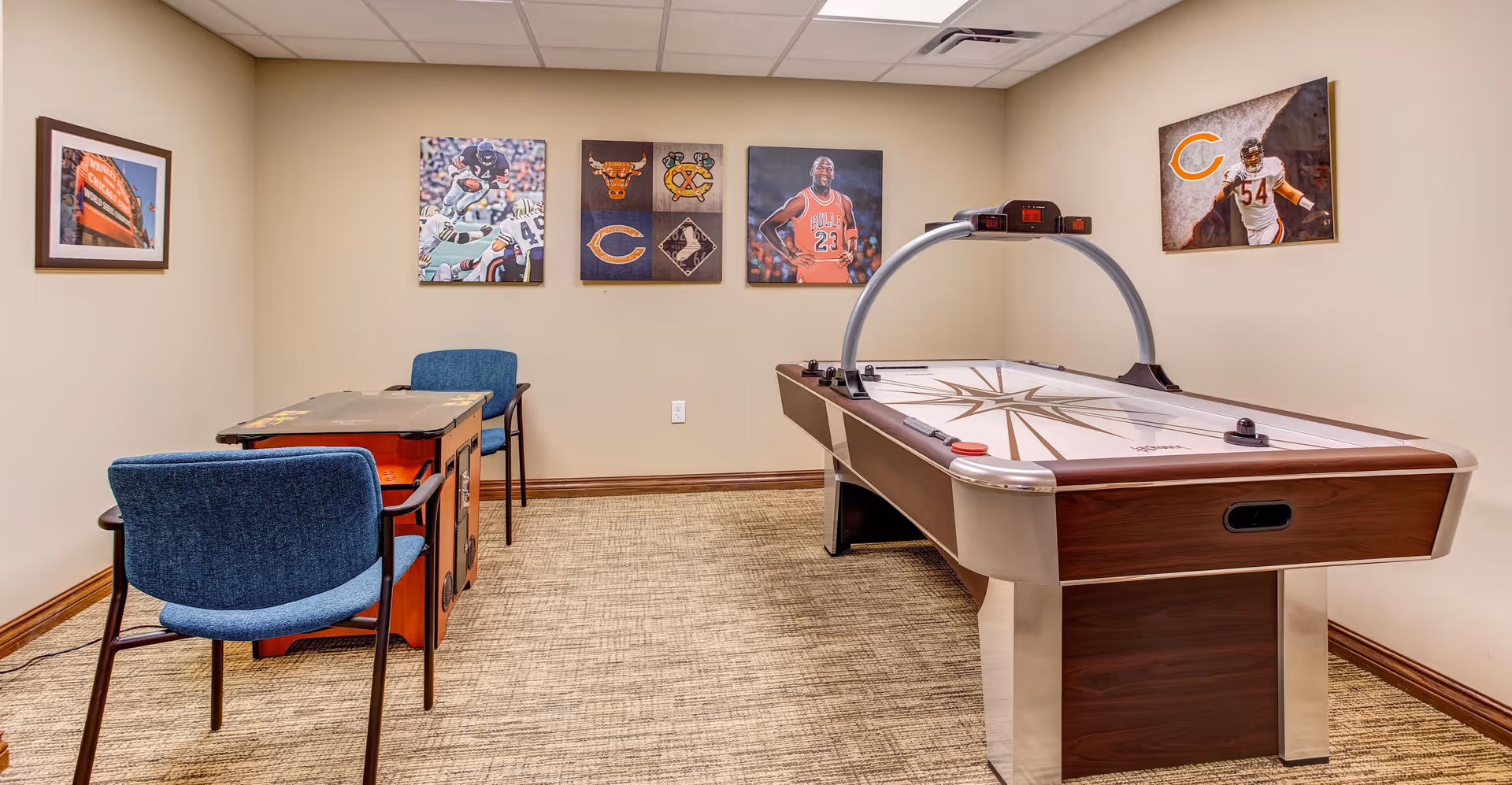 A game room with an air hockey table on the right and a vintage arcade game with two blue chairs on the left. The walls are decorated with framed sports-themed pictures, including images related to the Chicago Bears and Chicago Bulls.