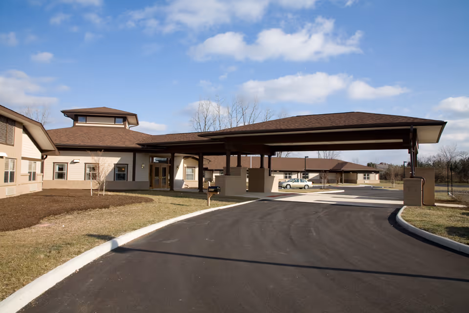 Exterior view of a single-story senior living facility building with a covered driveway entrance, a paved road, and a car parked near the building. The sky is partly cloudy and there is some grass and bare trees around the building.
