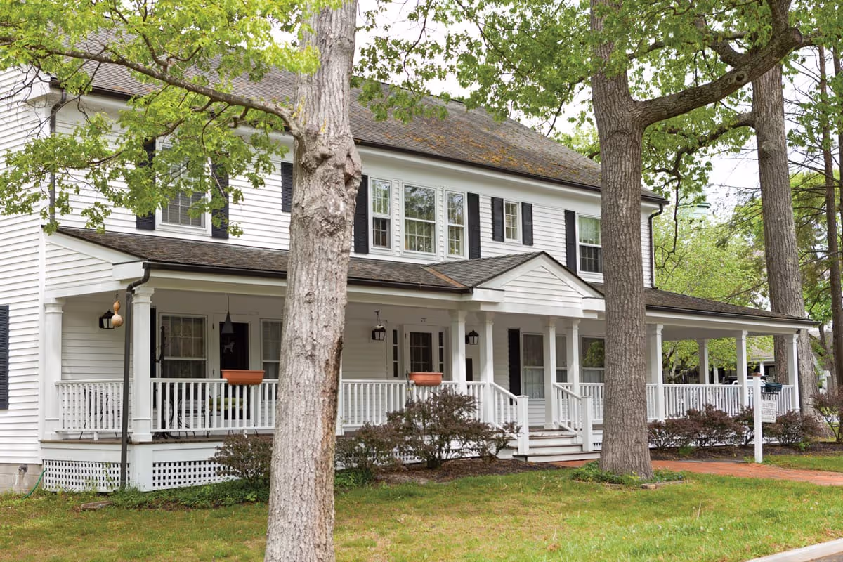 Two-story white colonial-style building with a wraparound porch and trees in the front yard.